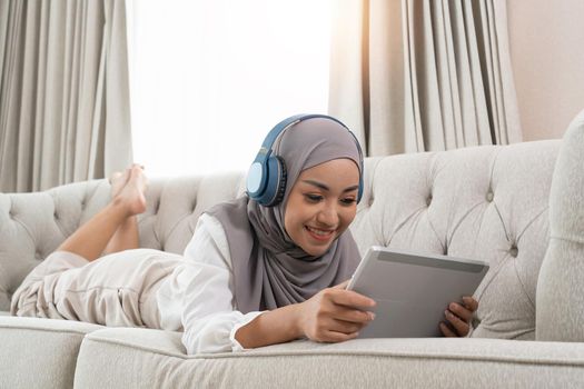 full length view of young muslim woman laying on couch in living room and watching movie on digital tablet