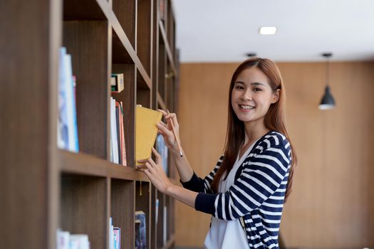 Asian female student stands in university library, looking for a book. Beautiful young woman in eyeglasses searching information for a project, learning for passing exams, education concept