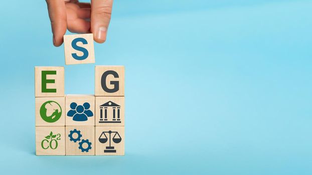 ESG concept of environmental, social and governance. Words ESG on a wood cubes. Hand holds wooden cube with abbreviation ESG standing with other icons on blue background