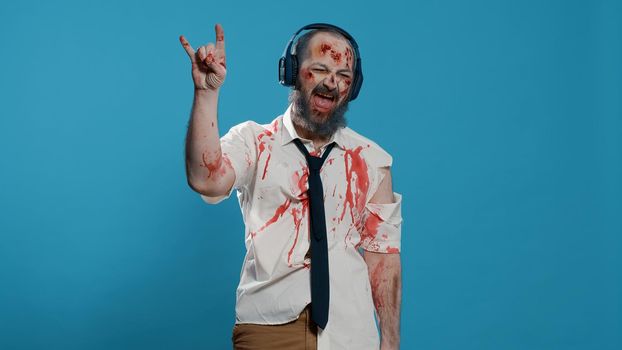 Messy creepy zombie wearing electronic wireless headphones while listening to music on blue background.