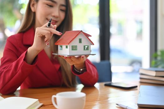 Smiling asian woman holding small house model. Mortgage and real estate investment concept.