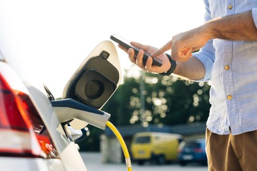Male attaching in power cord to electric car using app on smartphone. Man plugging electric car from charging station. Businessman charging electric car on charging station at sunset.