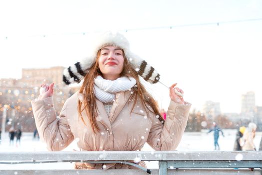 Medium shoot portrait of romantic European lady wears long stylish winter jacket and funny fluffy hat in snowy day. Outdoor photo of inspired blonde woman enjoying winter city.