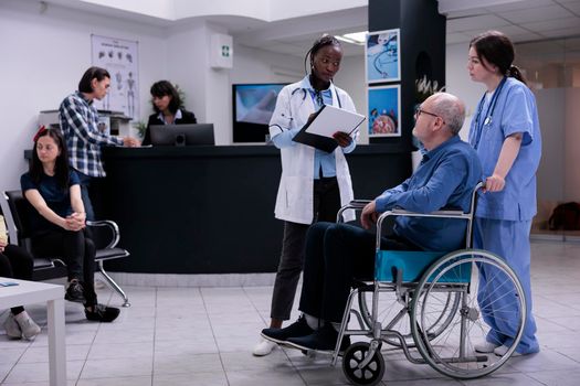 Older man living with disability being helped by caring nurse talking with doctor holding clipboard