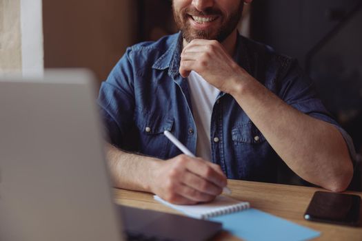 Hands of cheerful man with pen while taking notes at laptop and smiling. Male noting at computer.