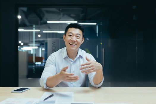 Online business training. Portrait of an active young Asian businessman. Conducts online business training on camera, sits at a table in a modern office, talks, explains, waves his hands, smiles