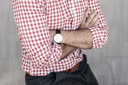 Torso of young entrepreneur wearing plaid shirt casualy leaning on desk with arms crossed against gray background wall.