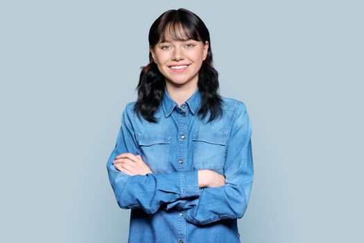 Portrait of beautiful teenage student woman looking at camera. Young charming brunette female smiling with teeth, confident with arms crossed on light gray color isolated background