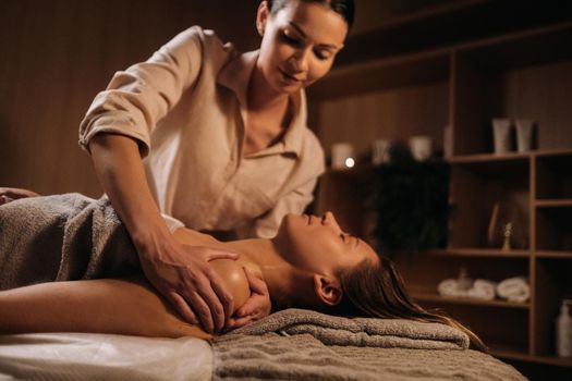 A masseuse gives a body massage to a woman in a spa center. A professional masseur massages the shoulder of a girl lying in a spa center