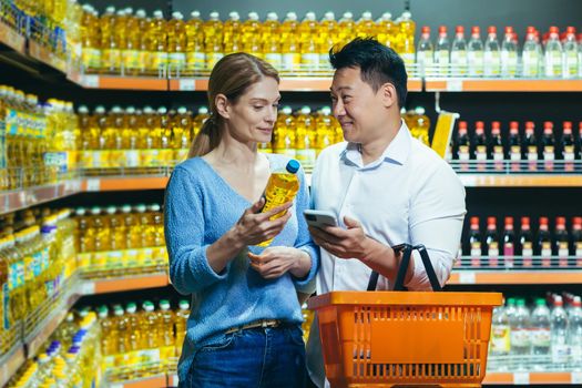Young Asian couple using smartphone in supermarket with shopping cart choosing picking products