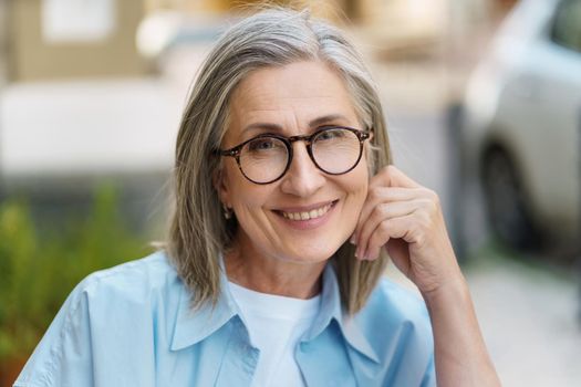 Confident charming mature woman smiling leaned head on hand wearing eye glasses and blue shit sitting outdoors with city background. Portrait of mature woman with grey hair and good skin