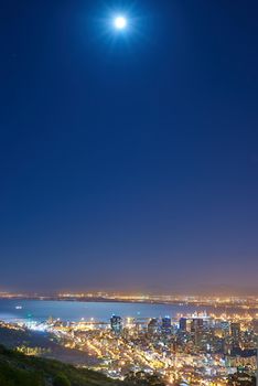 Urban city lights with a full moon in the midnight sky with copy space. Skyline with colorful lighting with the wide open ocean on the horizon. City buildings at night in Signal Hill, South Africa