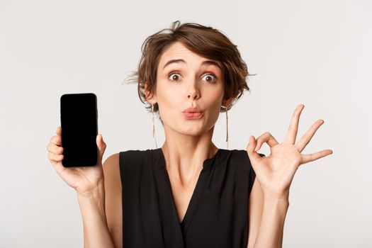 Amused pretty girl showing okay gesture and smartphone screen, standing over white background