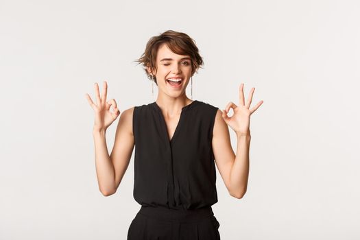 Sassy good-looking young woman assure you, showing okay gesture and winking, standing white background