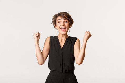 Excited successful young woman feeling lucky, smiling upbeat and fist pump, celebrating victory over white background