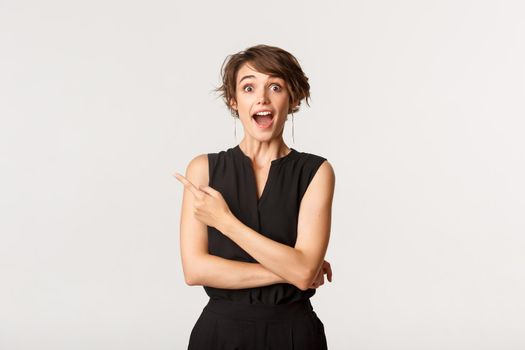 Portrait of happy and surprised caucasian woman open mouth fascinated, pointing upper left corner at promo banner, white background