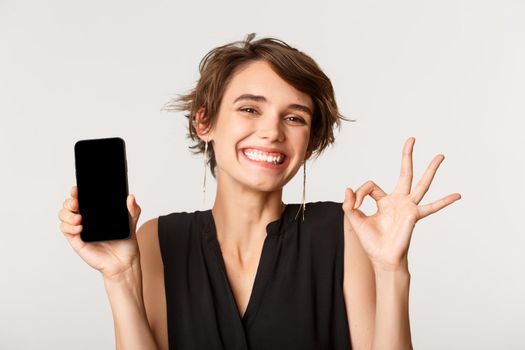 Image of cheerful pleased girl showing okay gesture, smiling and demonstrating mobile phone screen, standing over white background