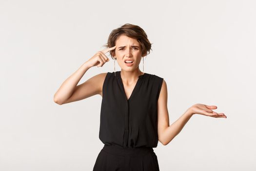 Image of frustrated woman scolding someone, pointing at head and shrugging bothered, standing white background