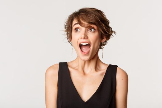 Close-up of excited, amazed young woman drop jaw and looking left, standing over white background