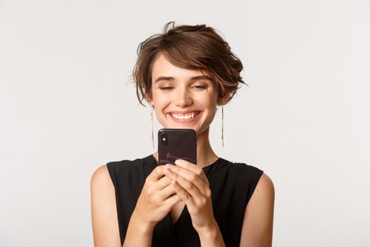 Close-up of satisfied young woman looking at mobile phone with pleased smile, standing over white background