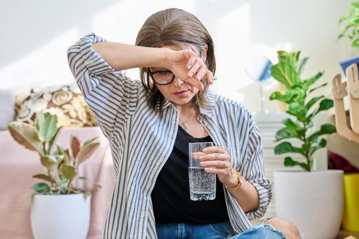 Mature woman suffering from headache, at home with a glass of water. Health, stress, depression, pain, migraine, older age problems