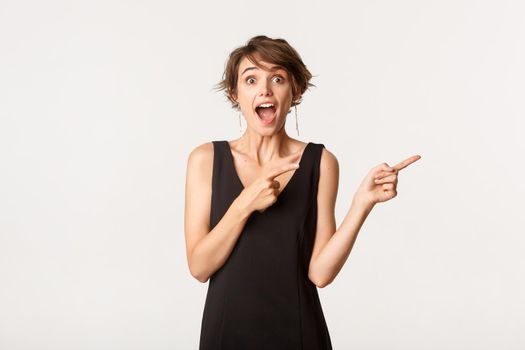 Image of surprised brunette woman looking amazed and pointing fingers right at logo, standing over white background