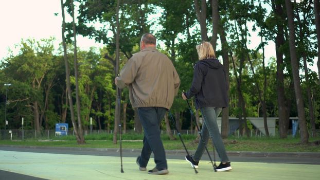 Pensioners are engaged in Nordic walking in the park. A man and a woman walk with sticks to improve health.