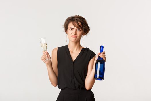 Skeptical and displeased young woman grimacing upset, holding bottle and glass of champagne, standing over white background