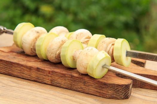 Zucchinis and mushrooms on metal skewers on wooden chopping board