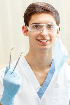 Portrait of handsome male dentist with stainless steel dental mirror and probe for tooth examining in the dental clinic. Doctor wearing white uniform, glasses and blue gloves