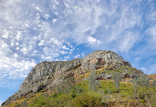 A photo of Lions Head and surroundings in Cape Town. Backdrop of rocky mountain with green vegetation and trees against blue sky filled with small wispy clouds. Barren landscape of natural wonder