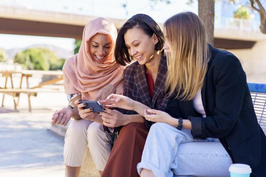 Positive multiethnic women sharing smartphone while resting on bench in park