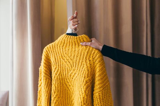 Close-up photo. The hand of a salesperson or consultant passing clothes, a yellow sweater on a hanger, to the hand of a client who is waiting in the fitting room behind the curtains. In a clothing store in a shopping center