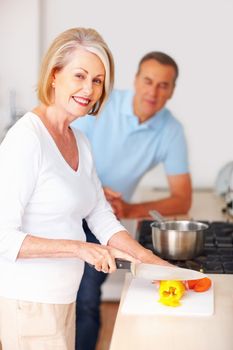 Smiling senior woman preparing food with man in background. Portrait of a smiling mature woman preparing food with man in background at kitchen.