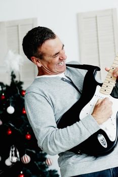 Mature man playing guitar with Christmas tree in background. Portrait of an energetic mature man playing guitar with Christmas tree in background.