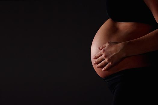 Pregnant woman feeling her baby against black - copyspace. Mid section of a pregnant woman feeling her baby against black background - copyspace.