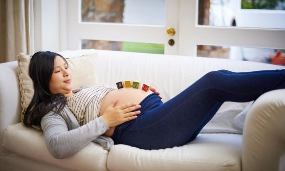 The beginning of something beautiful. an attractive young pregnant woman balancing wooden blocks on her tummy while relaxing on the sofa at home.