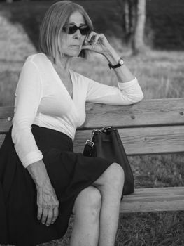 elegant caucasian senior woman wearing sunglasses sitting in a park at dusk