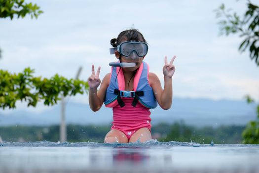 Cute little girl wearing goggles is sitting by the pool preparing for a diving lesson. Happy little girl is swimming and playing in outdoor swimming pool. Healthy Summer Activities for Kids.