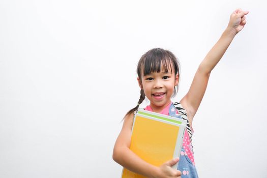 Back to school. Smiling little girl carrying a backpack holding books looking at the camera with arms raised on a white background with copy space. Girl glad ready to study.