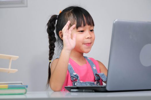 Smiling little Asian girl studying online having video call distant class with teacher using laptop. Happy little girl wave greeting with tutor while studying online with laptop at home. homeschooling
