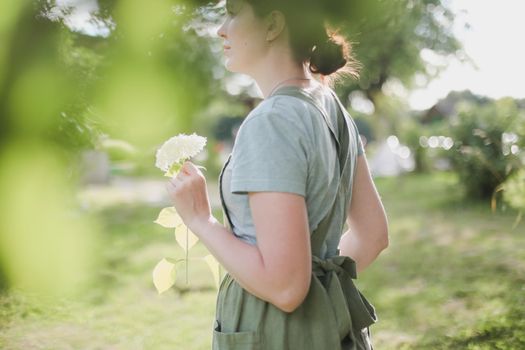 gardening and profession concept - young woman in apron holding flowers in the garden in summer