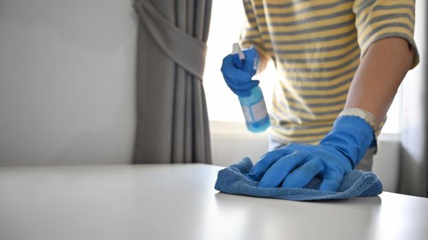 Young man holding disinfectant spray bottle and wiping dust on table with soft cloth.