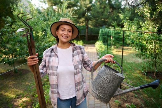 Charming Caucasian woman farmer with garden tools, smiles toothy smile looking at camera, while working in organic farm