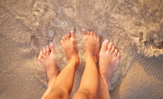Above shot of the feet of a caucasian couple in the ocean while standing on the sand at the beach. Romantic people sharing an intimate moment during a date by the seaside. Love and togetherness