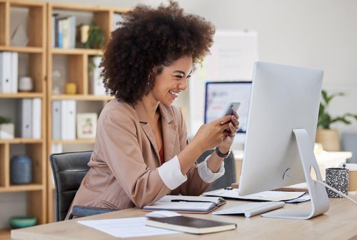 Happy young mixed race businesswoman with afro texting on cellphone while working on computer in an office. One female only checking social media and browsing apps online on smartphone to manage plans