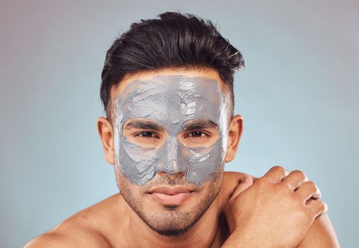 Portrait of one handsome young indian man applying an anti aging facial mask against a blue studio background. Mixed race guy wearing a moisturising clay or charcoal cream product on his face to get rid of blackheads for healthy, smooth and soft skin