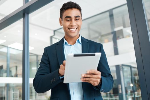 Young cheerful mixed race businessman holding and working on a digital tablet alone at work. One happy hispanic male businessperson smiling and using social media on a digital tablet in an office