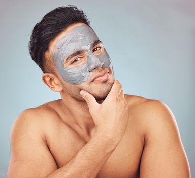 Portrait of one handsome young indian man applying an anti aging facial mask against a blue studio background. Mixed race guy wearing a moisturising clay or charcoal cream product on his face to get rid of blackheads for healthy, smooth and soft skin