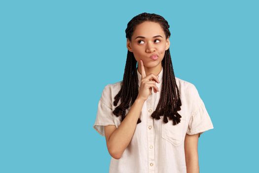 Woman with black dreadlocks keeps index finger on cheek, considers something, has pensive expression
